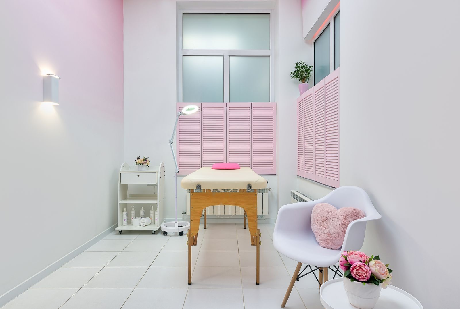 A bright treatment room with pink shutters, a treatment bed and fresh peonies